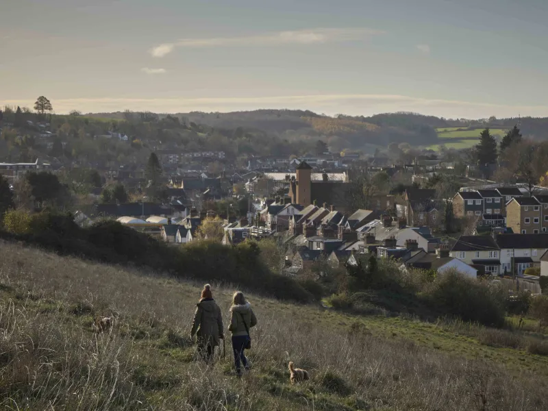 Two people walk a dog through fields. A town and rolling countryside are in the background.