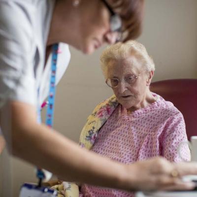 An elderly lady sitting in a chair on a hospital ward looks at a nurse as she takes notes