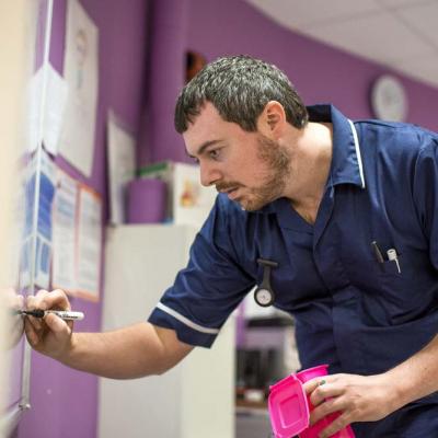 A matron in a dark blue uniform makes notes on a whiteboard on a hospital ward