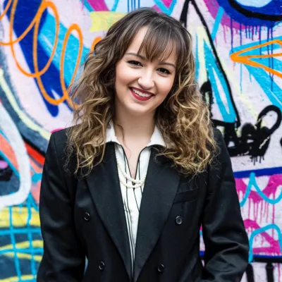 Headshot photo of Beth Kume-Holland, a woman in a smart suit jacket standing in front of a colourful graffitied wall