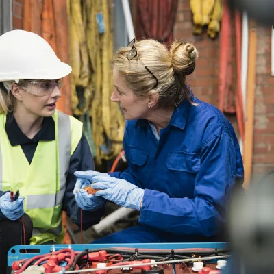 Two female mechanics repair machinery. The younger mechanic is dressed in a high-vis jacket, hard hat and safety goggles.