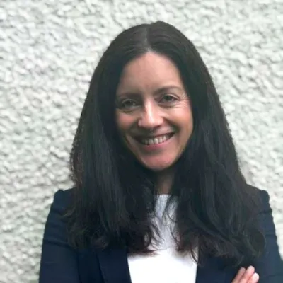 Alison Ismail, smiles with her hands crossed in front of a white background.