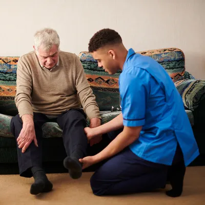 An image of a carer helping an older man stretch his legs at home. 